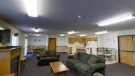 This single-floor space offers an open, functional layout that seamlessly integrates a living area, kitchen, and dining space into one cohesive environment. The living room is furnished with plush, dark green upholstered sofas and a wooden coffee table, creating a comfortable and inviting gathering space. Overhead, a textured ceiling with flush-mounted fluorescent lighting provides consistent, even illumination throughout the interior.

The fully equipped kitchen is positioned toward the rear of the open floor plan and features warm wood-toned cabinetry, a white refrigerator, oven range, and a dedicated countertop workspace — offering everything needed for meal preparation. A dining area with wooden chairs is situated adjacent to the kitchen, making it convenient for everyday dining.

The space also includes an interior door leading to a private room, adding to the overall utility of the layout. Neutral-toned walls paired with dark carpeting throughout contribute to a grounded, practical ambiance. A fire safety device is visibly mounted near the kitchen, reflecting attention to safety standards. Overall, this space is designed to be highly functional, accommodating both relaxation and day-to-day living within a straightforward, well-organized single-level footprint.