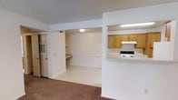 Located at 100 Chapelridge Dr in Richland, Mississippi, this single-story unit presents an open and functional layout designed for everyday living. The main living area features neutral-toned carpeting underfoot and a textured ceiling overhead, contributing to a clean, understated aesthetic throughout the space.

A half-wall partition separates the living area from the kitchen, creating an open-concept feel while maintaining a sense of defined space. The kitchen is equipped with warm oak-toned cabinetry, a white electric range with an overhead ventilation hood, and light-colored countertops. The pass-through design allows for easy interaction between the kitchen and main living space.

Adjacent to the living area, a built-in desk nook provides a dedicated workspace, complete with a wall-mounted shelf for storage and a built-in countertop surface. This area benefits from its own overhead lighting, making it practical for work or study. A door in the background leads to an additional room or entry area, adding further functionality to the overall floor plan. The consistent white walls throughout the unit create a bright and neutral backdrop, allowing for versatile interior styling.