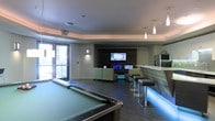 This single-floor entertainment lounge is thoughtfully designed to offer a comprehensive recreational experience. The space centers around a full-sized pool table finished in green felt, positioned prominently on dark hardwood-style flooring that runs throughout the room. Opposite the pool table is a comfortable lounge area furnished with contemporary green-toned sofas and chairs, arranged to face a feature wall housing a large flat-screen television flanked by additional screens, making it well-suited for watching sports or gaming. A stylish bar counter with integrated LED underlighting runs along one side of the room, accompanied by sleek bar stools with dark cushioned seats. Pendant lighting with elongated white fixtures hangs above the bar, complementing the recessed ceiling lights and wall-mounted sconces that provide layered illumination throughout. A slatted wood accent wall frames the media area, adding warmth and texture to the otherwise modern palette of whites and grays. Natural light filters in through a glass door on one side, offering a connection to the outdoors. The overall ambiance is contemporary, social, and versatile.