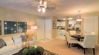 This single-floor residence showcases an open-concept design that integrates the living, dining, and kitchen areas into one cohesive, flowing space. The living area is finished with plush carpeting in a warm neutral tone and is illuminated by a ceiling fan with an integrated light fixture, providing both comfort and airflow. Decorative lamps add a soft, inviting ambiance, complementing the beige walls that carry consistently throughout the space.

Adjacent to the living area, a compact dining space accommodates a round table with upholstered chairs, making efficient use of the layout while maintaining a clean, functional aesthetic. The kitchen is positioned at the rear of the open floor plan and features white cabinetry, dark countertops, white appliances including a range and oven, and overhead lighting that brightens the workspace. Upper cabinets extend to the ceiling, maximizing storage capacity.

The entryway is defined by a white paneled door framed by neutral walls, offering a clear transition point into the main living space. Throughout, the warm color palette, consistent flooring, and coordinated lighting create a cohesive and comfortable atmosphere well-suited for everyday living.