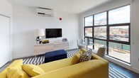 This single-floor studio offers a thoughtfully designed open-plan layout that efficiently combines living, sleeping, and cooking areas. The living space is anchored by a vibrant yellow sofa paired with a navy ottoman and a bold blue-and-white striped area rug, creating a lively, contemporary atmosphere. A wall-mounted split AC unit ensures year-round comfort, while large black-framed windows flood the space with natural light and provide views of the surrounding neighborhood.

The kitchen is sleek and functional, featuring slate-blue cabinetry, white subway tile backsplash, light countertops, and a full suite of stainless steel appliances including a refrigerator, dishwasher, range, and microwave. Open shelving above the counter provides additional storage and display space, complementing the modern aesthetic.

A sliding door closet offers built-in storage within the main living area, keeping the space organized and clutter-free. The bathroom is clean and crisp, finished in white with large-format gray floor tiles, a white floating vanity with under-cabinet storage, a well-lit mirror, and a tiled shower-tub combination. Dark hardwood-style flooring runs throughout the main living and kitchen areas, tying the space together with warmth and continuity.