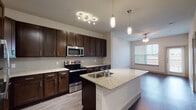 Located at 2250 Sara Jane Pkwy in Grand Prairie, Texas, this well-appointed single-floor residence combines contemporary finishes with a functional open-concept layout. The kitchen serves as the centerpiece of the living space, showcasing rich dark espresso cabinetry, light granite countertops, and a full suite of stainless steel appliances including a French door refrigerator, electric range, and built-in microwave. A subway tile backsplash adds a clean, modern touch, while pendant lighting above the granite-topped island — complete with a dual-basin sink — enhances both style and utility. Wood-look flooring flows throughout the main living areas, complementing the neutral palette and creating a warm, cohesive aesthetic. The open living area benefits from natural light streaming through large windows dressed with blinds, and a ceiling fan ensures year-round comfort. The primary bathroom features a spacious dual-sink vanity topped with granite, dark wood cabinetry, a large framed mirror, and wood-look flooring, with a walk-in closet visible beyond. The bedroom offers plush carpeting, a ceiling fan, and a generous window that brings in ample natural light. The overall design is cohesive and modern, with consistent dark wood finishes, granite surfaces, and neutral tones throughout.
