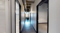 This urban loft showcases a striking industrial aesthetic across a single floor, thoughtfully designed to balance open communal spaces with private quarters. The open-concept living and kitchen area is the heart of the unit, featuring gleaming polished concrete floors that reflect abundant natural light pouring in through oversized windows. Exposed concrete beams and visible ductwork run across the ceiling, reinforcing the space's industrial character, while pendant lighting adds warmth over the centrally positioned kitchen island. The kitchen is outfitted with dark wood cabinetry, stainless steel appliances, and a large island with a stone countertop — ideal for both cooking and entertaining. A connecting hallway with the same polished concrete flooring and black-framed doors leads to the two bedrooms, each finished with neutral-toned carpet and clean grey walls, offering a quiet retreat from the open living areas. The primary bedroom provides a comfortable, well-proportioned space. The two bathrooms are finished to a high standard: one features a large glass-enclosed walk-in shower with white subway tile, a mosaic accent border, and a dark-vanity with an undermount sink; the second is finished with light tile flooring and a coordinating vanity. A dedicated closet completes the layout.