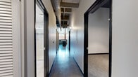 This urban loft showcases a striking industrial aesthetic across a single floor, thoughtfully designed to balance open communal spaces with private quarters. The open-concept living and kitchen area is the heart of the unit, featuring gleaming polished concrete floors that reflect abundant natural light pouring in through oversized windows. Exposed concrete beams and visible ductwork run across the ceiling, reinforcing the space's industrial character, while pendant lighting adds warmth over the centrally positioned kitchen island. The kitchen is outfitted with dark wood cabinetry, stainless steel appliances, and a large island with a stone countertop — ideal for both cooking and entertaining. A connecting hallway with the same polished concrete flooring and black-framed doors leads to the two bedrooms, each finished with neutral-toned carpet and clean grey walls, offering a quiet retreat from the open living areas. The primary bedroom provides a comfortable, well-proportioned space. The two bathrooms are finished to a high standard: one features a large glass-enclosed walk-in shower with white subway tile, a mosaic accent border, and a dark-vanity with an undermount sink; the second is finished with light tile flooring and a coordinating vanity. A dedicated closet completes the layout.