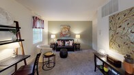 This single-floor residence offers a thoughtfully organized layout with distinct living, sleeping, dining, and kitchen areas. The living room is a welcoming space featuring soft carpeted flooring, neutral walls accented by a sage green feature wall, and natural light streaming through windows dressed with decorative valances and blinds. The room comfortably accommodates a seating arrangement and provides ample space for relaxed everyday living.

The bedroom is a quiet, carpeted retreat with a large window that brings in natural light filtered through stylish curtains. A wall-mounted TV and a built-in closet add practical convenience to the room's clean, simple design.

The kitchen is compact yet functional, equipped with wood-toned cabinetry, a range, and essential appliances to support everyday meal preparation. Adjacent to the kitchen, a dining or workspace area provides additional utility within the open floor plan.

The bathroom is efficiently designed with neutral tile flooring, a white vanity with a medicine cabinet and lighted mirror above, and a tub/shower combination finished with warm beige subway-style tile surround and chrome fixtures. Together, these spaces create a practical and comfortable single-floor living environment with a cohesive, understated aesthetic throughout.