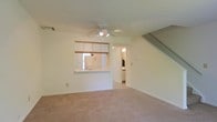 This two-floor space offers a functional and open layout designed for comfortable everyday living. The main level features a carpeted living area that provides a warm, understated foundation for the room. A ceiling fan with integrated lighting is centrally positioned overhead, offering both air circulation and ambient illumination throughout the space.

A defining feature of the main level is the pass-through kitchen window, which connects the kitchen to the living area, creating an open, communicative flow between the two spaces. The kitchen is equipped with white upper cabinetry trimmed with a wood accent rail, and a standard sink is visible beneath the pass-through counter. White appliances complement the clean, neutral color palette that carries throughout the home.

A doorway off the living area leads to an additional room, likely a bathroom or utility space, with visible lighting fixtures suggesting a functional, well-lit interior. A staircase with a simple railing rises along one wall, providing access to the upper floor and adding architectural dimension to the layout.

Overall, the space maintains a clean, neutral aesthetic with white walls, light-toned carpet, and straightforward finishes, making it a versatile and practical environment.
