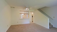 This two-floor space offers a functional and open layout designed for comfortable everyday living. The main level features a carpeted living area that provides a warm, understated foundation for the room. A ceiling fan with integrated lighting is centrally positioned overhead, offering both air circulation and ambient illumination throughout the space.

A defining feature of the main level is the pass-through kitchen window, which connects the kitchen to the living area, creating an open, communicative flow between the two spaces. The kitchen is equipped with white upper cabinetry trimmed with a wood accent rail, and a standard sink is visible beneath the pass-through counter. White appliances complement the clean, neutral color palette that carries throughout the home.

A doorway off the living area leads to an additional room, likely a bathroom or utility space, with visible lighting fixtures suggesting a functional, well-lit interior. A staircase with a simple railing rises along one wall, providing access to the upper floor and adding architectural dimension to the layout.

Overall, the space maintains a clean, neutral aesthetic with white walls, light-toned carpet, and straightforward finishes, making it a versatile and practical environment.