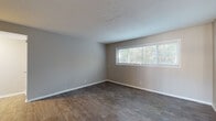 This single-floor home offers a well-organized layout across multiple rooms, as visible in the dollhouse view, with a practical and clean aesthetic throughout. The living areas feature durable dark hardwood-style flooring paired with neutral gray walls and textured ceilings, creating a calm and cohesive atmosphere. Wide horizontal windows allow generous natural light to fill these spaces, offering views of surrounding greenery. The kitchen is equipped with white cabinetry, a matching white range and dishwasher, a dark countertop with an integrated sink, and a tile backsplash. Light hardwood-style flooring and a sliding door with vertical blinds add brightness and outdoor access. The bedrooms vary in finish — some feature plush carpeting in warm tones alongside dual closets and ample wall space, while others showcase light hardwood-style flooring with built-in closet storage. Neutral beige and gray wall tones carry throughout the sleeping quarters, maintaining a consistent and versatile palette. White trim and baseboards provide clean contrast across all rooms. The overall ambiance is understated and move-in ready, offering a functional and adaptable living environment suited for a variety of furnishing styles.