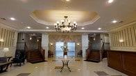 This grand lobby showcases a sophisticated architectural design centered around a striking multi-arm chandelier suspended from an octagonal tray ceiling with detailed crown molding. Recessed lighting complements the chandelier, ensuring the space is well-illuminated throughout. The focal point of the back wall is a floor-to-ceiling stone tile accent in warm beige and taupe tones, flanking two stainless steel elevator doors that provide a sleek, modern contrast to the classical surroundings. Symmetrical dual staircases with dark wood banisters and white balusters flank either side of the lobby, adding architectural grandeur and a sense of balance. The polished light-toned tile flooring reflects the ambient lighting, enhancing the spacious feel of the room. A decorative wrought-iron accent table centered in the space holds a vibrant floral arrangement, adding a welcoming touch. Wainscoting panels line the walls, reinforcing the traditional, upscale aesthetic. Wall sconces provide additional warm lighting throughout. Overall, the lobby exudes a timeless, elegant ambiance suitable for a distinguished residential or hospitality setting.