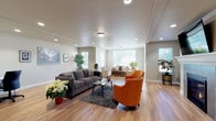 This single-floor communal lounge is thoughtfully designed to provide a warm and inviting gathering space. The open layout is anchored by light-toned hardwood flooring that flows seamlessly throughout, complemented by neutral, greige-toned walls that create a calm and comfortable atmosphere.

The room is organized into multiple distinct seating areas, offering flexibility for both intimate conversations and larger group gatherings. A central seating arrangement features a charcoal gray sofa paired with vibrant orange accent chairs atop a plush area rug, centered around a sleek coffee table. A secondary seating area in the background provides additional lounge space with neutral-toned furnishings.

A standout architectural feature is the tile-surround gas fireplace with a white mantel, above which a large wall-mounted flat-screen television is installed — creating a natural focal point for the room. Recessed lighting throughout the ceiling, supplemented by flush-mount fixtures, ensures the space is well-lit at all times.

Abundant natural light pours in through multiple large windows, offering views of lush outdoor greenery. Potted plants and floral arrangements are tastefully placed throughout, adding a lively, organic touch to the décor. A dedicated workstation with a desk and computer is also present, adding a practical element to this multifunctional space.