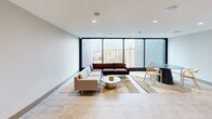 Located at 18545 Hilliard Blvd in Rocky River, Ohio, this modern communal lounge offers a thoughtfully designed, single-floor layout ideal for gathering and relaxation. The open-plan space is anchored by a generous seating area featuring a large sectional sofa and a stylish marble-topped coffee table, all arranged atop layered area rugs over light-toned flooring that runs throughout. Floor-to-ceiling windows along one wall flood the interior with natural light while offering views of the surrounding Rocky River cityscape, lending the room an open, airy character. Recessed ceiling lighting complements the natural light, ensuring the space remains bright and welcoming at any time of day. A casual dining nook with a round table and wooden chairs provides a secondary gathering area, while a built-in wooden shelving and storage unit along one wall adds both functionality and warmth, displaying books, games, and decorative objects. A potted indoor tree beside a glass partition wall introduces a touch of greenery, enhancing the inviting atmosphere. A closet provides convenient storage. The clean, neutral color palette—soft grays, warm taupes, and natural wood tones—gives the space a cohesive, contemporary feel well-suited for a community amenity room.