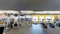 This single-floor fitness facility is designed to accommodate a wide range of workout needs in a spacious, functional environment. The gym features an open floor plan with durable rubber flooring throughout, providing both safety and comfort during exercise. The ceiling is finished with an industrial aesthetic, showcasing exposed ventilation ductwork and overhead fluorescent lighting that ensures the space is well-lit at all times. Large windows along the perimeter allow natural light to fill the room, contributing to an energizing atmosphere. The strength training area is outfitted with a variety of cable machines and free-weight equipment, including multiple Synergy-branded cable stations and squat racks, catering to both beginner and experienced lifters. The cardio zone is positioned near the windows and includes treadmills, ellipticals, and step machines, allowing users to exercise with a view of the outdoors. Bold yellow accent walls add a vibrant, motivating energy to the space. Mirrors are strategically placed to enhance the sense of openness and assist users with form during workouts. Overall, the facility is thoughtfully arranged to maximize usability while maintaining an open, inviting atmosphere suited for a variety of fitness routines.