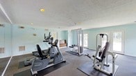 This fitness center offers a practical and well-organized workout environment designed to support a variety of exercise routines. The space is finished with light blue walls that lend a calm, refreshing atmosphere, complemented by a textured white ceiling with recessed lighting that ensures the room is well-lit throughout. Carpeted flooring with protective exercise mats beneath the equipment adds both comfort and functionality to the workout area. The equipment selection includes cardio machines such as a recumbent bike and an upright elliptical, alongside a multi-station cable machine and a dedicated strength training unit, providing users with options for both cardiovascular and resistance training. Natural light filters into the room through large windows and glass-paneled doors along one wall, creating an open and inviting feel. A small reception or check-in area is visible toward the rear of the space, along with a bulletin board, suggesting the facility is part of a larger managed property. The single-floor layout keeps everything accessible and easy to navigate, making this a convenient and straightforward space for daily fitness use.