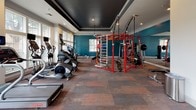 Located at 1717 E Belt Line Rd in Coppell, Texas, the St. Martin Fitness Center is a thoughtfully designed, single-floor facility built to support a comprehensive workout experience. The entrance features a tiled foyer with crown molding, neutral white walls, and a bold black-framed glass door that leads into the main workout space, setting a clean and welcoming tone from the moment you arrive.

The main fitness floor is spacious and well-appointed, featuring multi-toned patterned carpet throughout and a striking tray ceiling finished in deep charcoal, accented by recessed lighting and pendant fixtures. Bold teal accent walls add a dynamic visual energy to the space. Large multi-pane windows line multiple walls, flooding the interior with natural light and offering views of the surrounding outdoor greenery and community amenities.

The facility is fully equipped with a wide range of cardio equipment, including treadmills, StairMaster machines, ellipticals, and rowing machines — all positioned to take advantage of the natural light. A prominent red functional training rig anchors the center of the room, offering cable systems, suspension trainers, and versatile strength accessories. Full-length mirrors along one wall further enhance the sense of openness while supporting proper form during workouts. The overall ambiance is energetic yet refined, making it a well-rounded fitness destination.