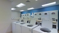 Located at 364 W Lane Ave in Columbus, Ohio, this self-serve laundromat offers a clean, well-maintained environment designed for convenience and efficiency. The single-floor facility operates 24 hours a day, making it accessible to customers at any time.

The main laundry area is outfitted with a generous lineup of commercial-grade top-load washing machines and stacked dryers, arranged in an orderly fashion to maximize usable space. The machines are modern and well-kept, providing customers with reliable performance for a variety of laundry loads.

The interior is bright and inviting, illuminated by recessed panel lighting set into a white drop ceiling that creates an evenly lit, comfortable atmosphere. A bold blue accent wall serves as a focal point, displaying the "Self-Serve Laundry" signage and reinforcing the facility's clean, professional aesthetic. Folding counters are conveniently positioned throughout the space, offering ample surface area for sorting and folding laundry. The overall design emphasizes functionality and cleanliness, with a crisp white-and-blue color scheme that feels fresh and hygienic. This laundromat is well-suited to serve the surrounding Columbus community with a practical, no-frills space built for ease of use.