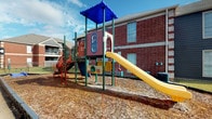 This community amenity facility is housed within a classic red brick building complex and offers a well-rounded set of shared outdoor and transitional spaces. The covered exterior corridor serves as a central access point, featuring white French doors, a smooth concrete floor, and a clean gray paneled wall. Vending machines and a wall-mounted emergency phone add practical convenience to this walkway, which is secured by a heavy-duty metal gate at its entrance.

Adjacent to the corridor, a spacious covered patio opens up to a beautifully maintained wooden deck supported by rich, dark-stained columns beneath a paneled ceiling. The deck overlooks a bright, resort-style swimming pool enclosed by ornamental iron fencing, with views extending to the surrounding residential buildings and lush landscaping. HVAC units and a pergola structure are visible on the deck level, adding to its functional character.

The outdoor grounds also include a dedicated children's playground, complete with a colorful multi-level play structure featuring a yellow slide, climbing elements, and interactive panels, all set on a wood chip safety surface beside the brick building exterior. The overall ambiance of the space is community-oriented, practical, and inviting, designed to offer residents a variety of outdoor recreational amenities in a well-maintained setting.