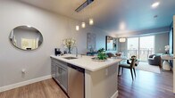 This contemporary single-floor residence is designed with clean lines, a neutral palette, and upscale finishes that create a cohesive and inviting atmosphere throughout. The open-concept kitchen serves as a central hub, anchored by a quartz-topped island with bar seating and pendant lighting overhead. Gray shaker-style cabinetry pairs elegantly with stainless steel appliances, including a full-size refrigerator, microwave, and dishwasher, while wood-look flooring ties the space together seamlessly into the living and dining area. The living room is bright and airy, benefiting from large floor-to-ceiling sliding glass doors that open to a private balcony, offering expansive outdoor views and an abundance of natural light. The bedroom is a serene retreat, featuring plush carpeting, generous natural light from a large window, and ample closet space. The bathroom is finished to a high standard, showcasing a dual-sink vanity with quartz countertops, gray shaker cabinetry, brushed nickel hardware, a large framed mirror, and a walk-in shower with subway tile surround. Wood-look flooring extends into the bathroom, reinforcing the unit's consistent, modern aesthetic. Overall, the space exudes a refined yet comfortable urban living experience.