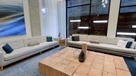 This sophisticated single-floor space makes a striking impression through its blend of raw industrial elements and refined modern design. Soaring ceilings amplify the sense of openness, while exposed concrete walls add an authentic, textural character that anchors the overall aesthetic. Floor-to-ceiling black-framed windows flood the interior with natural light and frame views of the surrounding environment, creating a seamless connection between indoors and out. A warm wood accent wall serves as a bold focal point, introducing natural warmth that balances the cooler concrete tones. The lounge area is thoughtfully appointed with a pair of clean-lined, light grey upholstered sofas accented with teal and neutral throw pillows, arranged around a substantial block-style coffee table crafted from natural reclaimed wood. A soft blue area rug grounds the seating arrangement, adding depth and color to the space. Decorative glass vases and subtle sculptural accents complement the curated, gallery-like atmosphere. Sheer white curtains soften the industrial framework of the windows, lending an airy elegance to the setting. Overall, the space strikes a refined balance between raw architectural materials and contemporary comfort, making it well-suited for social gatherings, casual meetings, or relaxed lounging.