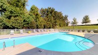 This outdoor pool facility offers a well-maintained swimming environment designed for relaxation and recreation. The centerpiece is a generously sized pool with clear, blue water, featuring clearly marked shallow and deeper sections for safety and convenience. The surrounding concrete deck provides ample space for lounging, lined with multiple chairs and a shaded umbrella seating area along the perimeter fence, making it ideal for extended outdoor enjoyment.

A white metal fence encloses the pool area, providing both safety and a clean, classic aesthetic. Tall, mature trees border the space on multiple sides, creating a natural green backdrop that offers a sense of seclusion and shade. Open lawn areas extend beyond the fence, adding to the expansive outdoor feel under a broad, open sky.

Access to the facility is provided through a secured brick entryway, featuring a wall-mounted intercom system and an information panel alongside a glass-paned door — reinforcing a controlled, community-oriented environment. The entry structure is built from classic red brick, consistent with the surrounding architectural character.

An additional interior brick-walled area with a concrete floor appears to serve as a utility or support space for the pool facility, equipped with electrical outlets and open framing, suggesting functional infrastructure behind the scenes.