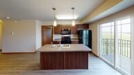 Located at 3450 38th Ave S in Fargo, North Dakota, this single-floor unit offers a well-designed, open-concept layout with durable luxury vinyl plank flooring throughout the main living areas. The kitchen serves as the centerpiece of the space, featuring rich dark wood cabinetry, a large central island with a built-in sink and pendant lighting overhead, and a full suite of black appliances including an electric range, over-the-range microwave, and refrigerator. A sliding glass door adjacent to the kitchen invites natural light and provides outdoor access. The kitchen opens seamlessly into a spacious living area, which includes a dedicated built-in desk nook with shelving — ideal for a home office setup. The bedrooms are carpeted for comfort and include generous walk-in closet space outfitted with wire shelving systems. Two bathrooms serve the unit: one features a dark wood vanity with tile flooring, while the second offers a single-sink vanity with a tub-shower combination. A dedicated laundry area houses a full-size Whirlpool washer and dryer, adding significant convenience. Windows throughout provide views of the surrounding landscape and keep the space bright and airy.