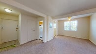 This single-floor residence offers a straightforward and functional layout, as illustrated by the overhead floor plan view. The living area is generously sized, finished with neutral wall-to-wall carpeting and painted in a soft cream tone that gives the space a clean, understated appearance. A ceiling fan provides air circulation, and a wide window fills the room with natural light while offering views of surrounding greenery.

Adjacent to the living area, the kitchen is practical and well-equipped, featuring warm honey-toned wood cabinetry that extends both above and below the countertops. A stainless single-basin sink, a gas range with an overhead ventilation hood, and a full-size refrigerator complete the kitchen's functionality. The floor is finished with light-toned tile, providing an easy-to-clean contrast to the carpeted areas.

The bedroom is a comfortable, well-proportioned room with plush carpeting, a ceiling fan, a wall-mounted HVAC unit, and a window that invites natural light. A secondary room or flex space is also present, similarly carpeted and bright.

The bathroom includes a full bathtub with tile surround, mosaic tile flooring, a toilet, a vanity with sink, and a window fitted with blinds for privacy and natural ventilation.