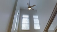 This two-story space blends rustic charm with modern sensibility, offering a distinctive character defined by its architectural details. Exposed wooden ceiling beams and warm hardwood flooring establish a rich, textural aesthetic throughout the interior, evoking a classic loft-style atmosphere. Flush-mount ceiling lights and wall sconces provide consistent, well-distributed illumination that complements the natural warmth of the wood elements.

Moving through the space, a hallway connects the main areas with a sense of flow and purpose, leading toward a striking open room with dramatically tall ceilings. This expansive area draws the eye upward to soaring walls fitted with large windows adorned with horizontal blinds, inviting generous amounts of natural light while maintaining privacy. A ceiling fan is centrally positioned overhead, ensuring comfort across seasons.

The staircase connecting the two floors is visible from the main living area, reinforcing the vertical scale of the space and contributing to its open, airy feel. The overall ambiance is one of understated character — a thoughtful balance between the raw, historic texture of exposed beams and reclaimed-style wood and the clean, bright neutrals of the upper living areas. This space is well-suited for those who appreciate architectural depth and a sense of volume in their environment.
