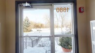 This single-floor building presents a practical and functional entryway design. The main entrance is framed by a set of glass doors prominently displaying the address "1880," allowing natural light to filter into the interior lobby. The glass panels offer views of the surrounding winter landscape, characterized by snow-covered grounds, mature evergreen and deciduous trees, and an open parking area beyond the steps.

Exterior access is provided via a set of steps flanked by metal railings, with a potted evergreen placed near the entrance as a welcoming touch. The entry vestibule features warm beige-toned walls and is equipped with a fire alarm panel, reflecting standard safety provisions. A built-in bank of numbered metal mailboxes lines one wall of the entryway, providing residents with convenient, secure mail access directly upon entry.

The overall aesthetic is clean and utilitarian, with neutral tones and durable finishes suited for a multi-unit building. The glass-forward entrance design ensures the lobby remains bright even during winter months, connecting the interior to the natural surroundings outside.