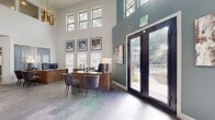 This single-floor commercial space presents a thoughtfully designed interior that blends contemporary style with functional elegance. The reception area commands immediate attention with its soaring double-height ceilings lined with a row of clerestory windows that flood the space with natural light. A sleek, warm walnut reception desk anchors the area, accompanied by plush gray guest chairs, while a striking gold chandelier adds a refined accent overhead. Dark-framed French doors at the entrance offer views of lush greenery outside, creating a welcoming first impression.

Adjacent to the reception, an open lounge area features a sophisticated teal accent wall, neutral upholstered sofas, a tufted leather bench coffee table, and layered area rugs atop large-format tile flooring. Curated artwork and symmetrically arranged table lamps lend a polished, gallery-like quality to the space. Recessed lighting and crown molding further elevate the interior's refined aesthetic.

A connected meeting and dining area offers a round table with curved upholstered chairs, bathed in natural light from multiple windows dressed with clean white blinds. A dark storage credenza and decorative accessories complete the space, making it well-suited for both casual gatherings and professional meetings. Throughout, the consistent use of soft neutrals, teal accents, and natural textures creates a cohesive and inviting atmosphere.