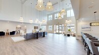 Located at 18183 Wheeler Rd in Westfield, Indiana, this single-floor community space offers a generously sized, thoughtfully designed interior that blends modern aesthetics with welcoming functionality. The centerpiece is a grand open living area distinguished by dramatic double-height ceilings and an impressive array of pendant lighting in varied woven and globe styles, casting a warm, layered glow throughout the space. Light hardwood-style flooring runs continuously underfoot, complementing the crisp white walls and creating a bright, airy atmosphere. Expansive windows and glass doors along the perimeter flood the interior with natural light and provide views of the surrounding outdoor areas. Multiple seating arrangements — including plush sofas, accent chairs, and intimate conversation clusters — offer flexible gathering spaces for a variety of uses. The kitchen features a large central island with bar seating, white shaker cabinetry, granite countertops, a subway tile backsplash set against a bold slate-blue accent wall, stainless steel appliances, and stylish rattan pendant lights. The dedicated dining room accommodates a substantial dark-wood dining table with upholstered seating, accented by contemporary landscape artwork and a glass door leading directly to the outdoors. A quiet lounge area with large windows rounds out this well-appointed space.