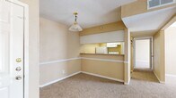 This single-floor residence offers a straightforward and functional layout designed for comfortable everyday living. Warm beige tones carry throughout the space, complemented by plush wall-to-wall carpeting that adds a soft, cohesive feel to the living, dining, and bedroom areas.

The dining area is anchored by a classic pendant light fixture and features a built-in pass-through window that opens directly to the kitchen, creating a convenient connection between the two spaces. The kitchen is equipped with white appliances, cabinetry, and mirrored upper accents that reflect light and add visual depth. A hardwood-accented entry near the front door provides a welcoming transition into the main living space.

The living room is generously proportioned and benefits from a large sliding glass door fitted with vertical blinds, allowing abundant natural light to fill the room while offering access to the outdoors. A secondary bedroom features a standard window with blinds, providing privacy and additional light.

The bathroom is clean and functional, outfitted with a full tub and shower combination, tiled surround, and standard fixtures. White trim and chair-rail molding detail the walls in key areas, adding a touch of classic character. The overall ambiance of the space is bright, neutral, and move-in ready.