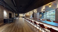 This expansive single-floor amenity space is designed with an upscale, industrial-chic aesthetic that blends warm wood tones, exposed brick, dark ceilings, and metal accents throughout. The layout is thoughtfully zoned into several distinct areas, each with its own character while maintaining visual cohesion.

A long bar counter runs along one wall, lined with rows of cognac-colored leather bar stools and illuminated from below by integrated lighting, creating a striking hospitality-style focal point. Adjacent to the bar, a kitchenette alcove features stainless steel appliances framed by richly toned metallic tile walls and herringbone brick flooring.

The game area anchors the central zone with a full-size billiards table beneath a sculptural pendant light, accompanied by a foosball table. Nearby, a dramatic accent wall constructed from circular wood medallions adds artistic texture, visible through steel-framed glass partitions that define the space without fully enclosing it.

A dedicated screening and lounge area offers plush teal velvet armchairs and a generous sectional sofa arranged before a large-format wall-mounted display, surrounded by dark wood paneling and dramatic drapery. Additional lounge nooks feature exposed brick walls, curated artwork, and light wood flooring, contributing to the space's refined yet relaxed atmosphere.
