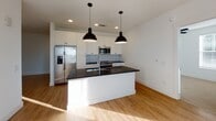 This thoughtfully designed single-floor residence showcases a clean, contemporary aesthetic with high-quality finishes carried consistently throughout the space. The open-concept kitchen serves as a central highlight, featuring white shaker cabinetry extending to the ceiling, a striking dark granite countertop island with an integrated sink, stainless steel appliances, and a herringbone-patterned tile backsplash. Two bold black pendant lights anchor the kitchen visually, while recessed lighting and warm hardwood-style flooring add to the bright, inviting atmosphere. Adjacent to the kitchen, a dedicated wet bar area includes a built-in wine cooler, open shelving with a wooden accent shelf, and matching dark countertops — an ideal space for entertaining. The bedrooms are carpeted in a neutral tone, offering comfort and warmth, with large windows fitted with blinds that invite generous natural light and outdoor views. A ceiling fan enhances comfort in one of the bedrooms. The bathrooms are well-appointed, with one featuring a double vanity topped with dark granite, dual black-framed mirrors, wall-mounted sconce lighting, and a tub-shower combo with vertical tile surround. Gray shaker-style interior doors with matte black hardware provide a cohesive, modern detail throughout the hallways and rooms.