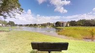 This single-floor residence is situated within a well-maintained community that boasts a tranquil, park-like setting. The most striking feature of the property is its access to stunning waterfront views, where a scenic pond takes center stage, complete with a decorative fountain that adds a touch of elegance to the natural landscape. Lush, manicured green lawns stretch along the water's edge, complemented by mature trees that provide shade and privacy. A classic wooden bench positioned along the lakefront invites residents to sit and enjoy the peaceful outdoor surroundings beneath open, cloud-dotted skies — a hallmark of a warm Florida-style climate. The broader community setting includes multi-story residential buildings visible across the water, reinforcing the neighborhood's established and cohesive character. The exterior environment reflects a thoughtfully designed community with an emphasis on outdoor living and natural beauty. While interior images were limited in visibility, the overall setting suggests a residence that places significant value on its connection to the outdoors, offering a serene retreat with picturesque water and landscape views as a daily backdrop.