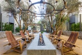Located at 27665 Forbes Rd in Laguna Niguel, California, this single-level outdoor courtyard space offers a thoughtfully designed amenity area suited to Southern California's year-round climate. The space is anchored by a striking pergola structure featuring reclaimed wood beams and rope-hung Edison bulb pendants, casting warm ambient light over the stone-paved entertaining area below.

Beneath the pergola, a built-in outdoor kitchen is equipped with stainless steel appliances, a marble-topped counter, and a utility sink, providing a fully functional preparation area for outdoor gatherings. A standout feature of the courtyard is a decorative mosaic-tiled wood-fired pizza oven, serving as both a functional cooking element and an artistic focal point. Natural stone paving extends throughout the space, lending a cohesive and upscale aesthetic.

The courtyard is generously landscaped with mature olive trees planted in raised stone-clad planters, complemented by agave, tropical foliage, and low-water Mediterranean plantings. Wooden dining tables and woven chairs provide comfortable seating throughout. A gated arched entryway framed by tall palm trees and dark stone masonry creates a grand sense of arrival, connecting the outdoor space to the surrounding property with architectural elegance.
