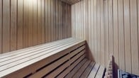 This dry sauna is constructed entirely with light-toned, vertically slatted natural wood paneling that covers every surface — walls, ceiling, and floor — creating a warm, cohesive aesthetic consistent with traditional sauna design. The interior is compact yet functional, with slatted wooden benches providing comfortable seating or reclining space during sessions. The bench construction follows classic sauna conventions, with open slats beneath the seating platform to allow for proper air circulation and heat distribution throughout the space.

A dedicated rock sauna heater is positioned at floor level, loaded with volcanic stones that absorb and radiate dry heat evenly into the room. This setup is central to the sauna's function, enabling the high, consistent temperatures characteristic of a dry sauna experience. Subtle recessed lighting provides a soft, ambient glow, enhancing the calming atmosphere without disrupting the natural warmth of the wood tones.

The entry features a solid wood door with a small glass window, allowing for visibility into the space while maintaining heat retention. Usage guidelines are posted on the interior wall, ensuring a safe and informed experience for users. Overall, the sauna offers a clean, traditional, and well-appointed environment designed for relaxation and wellness.