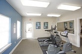 This fitness center is a compact yet fully functional workout space designed to support a well-rounded exercise routine. The room features wall-to-wall carpeting underfoot and is illuminated by overhead fluorescent lighting, creating a bright and functional environment. Two distinct color zones define the space — warm beige tones on one side transition into a teal accent wall on the opposite end, adding visual interest to the interior.

The cardio section includes a treadmill, an elliptical trainer, and a stationary recumbent bike, each positioned to allow comfortable use without overcrowding. For strength training, a multi-function cable machine serves as the centerpiece, accompanied by a weight bench and leg extension equipment, offering options for both upper and lower body workouts. Instructional fitness posters are displayed on the walls, serving as handy exercise references.

Large mirrors are mounted along one wall, expanding the perceived depth of the room and allowing users to monitor their form. Multiple windows with blinds line the teal-accented wall, introducing natural light while maintaining privacy. Two exterior doors provide convenient access points, with one offering a glass panel view to an outdoor walkway. The overall layout is efficiently organized, maximizing the use of available space.