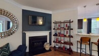 This single-floor home in Cary, NC offers a thoughtfully designed layout with distinct spaces that blend functionality and modern style. The living room serves as a focal point, featuring a classic white-mantled fireplace set against a bold deep-blue accent wall with a wall-mounted television above. An open-style shelving unit with a wood-and-metal frame adds a contemporary touch. Crown molding and warm hardwood flooring run throughout the main living areas, lending a polished finish.

The dining area accommodates a round wood table surrounded by vibrant red chairs, bathed in natural light from large windows dressed with patterned dark curtains. The galley-style kitchen is well-equipped with dark espresso cabinetry, granite countertops, a tile backsplash, and stainless steel appliances including a refrigerator, range, and microwave.

The bedroom features a deep green accent wall, carpeted flooring, a ceiling fan, and a generous window with outdoor views. Bathrooms include one with a sliding glass-door shower and wood-look flooring, and another with a tiled tub-shower combination. A dedicated laundry area houses a full-size washer and dryer with overhead shelving for added storage.