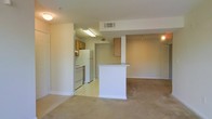 This single-floor space features an open-concept design that seamlessly connects the kitchen and living area. The kitchen is equipped with medium-toned wood cabinetry, a white refrigerator, and a stove, with light-toned vinyl flooring that defines the cooking zone. A white breakfast bar serves as a natural divider between the kitchen and the carpeted living area, providing both additional counter space and a casual dining or workspace option. The living area is finished with neutral wall-to-wall carpeting, contributing to a warm, understated atmosphere. Overhead, a ceiling vent ensures climate control throughout the space. A doorway visible beyond the kitchen hints at additional rooms, likely including a bathroom or bedroom. The overall color palette is neutral, with white walls and trim, creating a clean and adaptable backdrop. Recessed and flush-mount lighting fixtures provide consistent illumination throughout. The layout is efficient and functional, making good use of the available floor space across both zones.