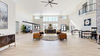 Located at 3555 Sirocco Way in Lafayette, Indiana, this expansive single-floor community clubhouse offers an impressive and thoughtfully designed gathering space. The centerpiece is a grand open living area with soaring double-height ceilings, flooded with natural light from a sweeping bank of clerestory and large-pane windows. Oversized ceiling fans overhead and light-toned hardwood-style flooring throughout lend a clean, contemporary feel to the entire space.

The main lounge area features a stylish seating arrangement of cognac leather sofas and chairs anchored by a bold circular area rug, creating an inviting focal point. A dark, gold-accented reception or concierge counter adds an elegant contrast against the bright surroundings. Nearby, an exposed brick accent wall serves as a dramatic backdrop for a built-in beverage and coffee bar, complete with beverage coolers and counter seating — ideal for social gatherings. A dining area with tables and stools complements this amenity zone.

A separate, generously sized multipurpose room provides flexible open space, while a dedicated game room features classic arcade machines for recreational use. A kitchen and bar area with counter seating rounds out the amenities. Staircase access and an upper-level overlook further enhance the architectural character of this vibrant, community-focused space.