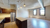This single-floor urban loft showcases a compelling blend of industrial character and modern finishes throughout its open layout. The living and kitchen areas flow seamlessly together, anchored by striking architectural details including exposed silver ductwork, painted corrugated ceilings, and prominent decorative columns that speak to the building's converted heritage.

The kitchen is a standout feature, offering rich dark-walnut cabinetry, light-toned countertops, and a full suite of stainless steel appliances including a refrigerator, range, and built-in microwave. A central island provides additional prep and seating space, while pendant lighting overhead adds warmth and ambiance. Track lighting runs along the ceiling, providing flexible illumination across the open-plan space.

The generously proportioned living area is bathed in natural light from large, black-framed windows that offer views of the surrounding cityscape and skyline. Light hardwood flooring runs throughout, complementing the bright, airy atmosphere.

The bathroom is finished with a clean, contemporary aesthetic, featuring large-format tile flooring, a vanity with dark countertop and under-mount sink, and a walk-in shower enclosed by sliding glass doors with tiled surround. The overall ambiance of this loft is one of industrial elegance — spacious, light-filled, and thoughtfully designed.