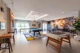 This single-floor amenity lounge is thoughtfully designed to blend comfort, style, and entertainment in one expansive, open-plan space. The main lounge area is anchored by a sleek, light blue channeled sofa paired with coordinating armchairs, all arranged atop a bold geometric black-and-white area rug over warm hardwood flooring. A large wall-mounted flat-screen TV sits above a linear gas fireplace set into a dramatic dark accent wall, creating a sophisticated media focal point. Ambient cove lighting runs along the tray ceiling, casting a warm glow throughout the space. A striking multicolored textile wall hanging adds an artistic pop of color to the lounge area. Structural white columns define zones within the open floor plan while a slatted wood ceiling detail transitions the space into the games and recreation area beyond. This zone features a foosball table, a pool table, and a dedicated dining or game table, all bathed in natural light from large windows. A decorative open wood shelving unit filled with books, plants, and curated objects adds warmth and visual interest. The overall ambiance is modern and inviting, combining curated artwork, rich wood tones, and vibrant accent pieces to create a dynamic communal environment.