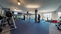 This expansive fitness center is designed to accommodate a full range of workout styles within a single, well-organized floor. The space is defined by its open layout, dark navy flooring, and a bright, airy atmosphere created by floor-to-ceiling windows that allow generous natural light and offer views of the surrounding outdoor environment. The cardio zone is lined with a row of treadmills and elliptical machines, each equipped with individual screens, and is positioned directly in front of the large windows to provide an energizing workout environment. Full-length mirrors along the walls amplify the sense of space and are a practical feature throughout. The strength training area is generously appointed with a power rack, cable machines, a dedicated dumbbell station, a heavy punching bag, and a functional training rig with battle ropes. A standout design element is the bold steampunk-themed mural—featuring oversized clock faces and intricate gear motifs—that spans entire walls in the weight training section, giving the space a distinctive and dynamic character. Additional cardio equipment, including stationary bikes and stair climbers, rounds out the offering. The entry area provides a welcoming transition with a coffered ceiling detail, built-in wooden cubbies, and decorative artwork, setting an organized and polished tone for the facility.