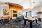 This thoughtfully designed single-floor amenity space seamlessly blends social, recreational, and functional areas into one cohesive environment. The heart of the space is a generously sized open lounge anchored by a stone-surround gas fireplace and a wall-mounted television, creating a warm and inviting focal point. Above, a dramatic oversized chandelier crafted from rope and cascading Edison bulbs serves as a striking artistic centerpiece, complemented by pendant globe lights throughout.

The game area features a full-size pool table positioned beneath warm pendant lighting, with cue storage conveniently nearby, and a dartboard station tucked along the wall. Comfortable curved sofas and accent chairs are arranged throughout the lounge, encouraging relaxed conversation or casual gatherings.

Natural light pours in through large sheer-curtained windows and glass French doors that open to an outdoor pool area, creating a bright and airy atmosphere. An abstract-patterned area rug grounds the carpeted lounge space, adding depth and texture underfoot.

A separate dining and kitchenette area transitions from carpet to rich hardwood-style flooring, featuring white cabinetry, a sink, countertop appliances, a wine/beverage refrigerator, and a dining table with seating for a group. Nature-inspired artwork and lush greenery add personality and warmth throughout the entire space.