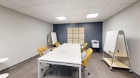 Located at 25 Winooski Falls Way in Winooski, Vermont, this single-room meeting space offers a well-appointed environment designed for focused collaboration and small group gatherings. The room features a sleek white rectangular conference table at its center, surrounded by cushioned golden-yellow rolling chairs accented with black-and-white houndstooth pillows, providing both comfort and style during extended sessions. Two rolling whiteboards are positioned within easy reach, supporting brainstorming and presentations. A standout design element is the deep navy accent wall adorned with a large typographic art installation featuring an F. Scott Fitzgerald quote, adding an inspirational and literary character to the space. The remaining walls are finished in a neutral light gray, creating a clean, professional backdrop. Flooring throughout is a warm wood-tone laminate, lending a contemporary yet inviting feel. A cozy lounge corner near the glass-panel entry door features a colorful geometric-print accent chair and a small round side table, offering a casual seating option. The drop ceiling is fitted with recessed LED panel lights that provide bright, even illumination. A multifunction printer and fire extinguisher are also present, reflecting practical workplace readiness. The glass door entry connects the room to a larger commercial corridor, ensuring easy accessibility.