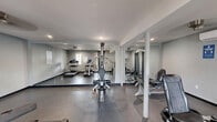 Located at 8003 SW Benaroya Lane in Huntsville, Alabama, this dedicated fitness center offers a practical and efficiently organized exercise space on a single floor. The room is finished with neutral gray walls, durable rubberized flooring, and recessed lighting complemented by a ceiling fan, creating a clean and functional workout environment. A wall-mounted mini-split HVAC unit ensures climate control year-round, keeping the space comfortable during all seasons. Large floor-to-ceiling mirrors line one wall, visually expanding the room and allowing users to monitor their form during exercise. The cardio section is well-appointed with multiple treadmills equipped with digital displays, as well as an elliptical trainer, catering to a variety of aerobic workout preferences. A multi-function cable machine serves as the centerpiece for strength training, accompanied by additional weight and resistance equipment. Multiple windows allow natural light to filter into the space, enhancing the overall atmosphere, while a glass exterior door provides direct outdoor access and adds to the sense of openness. The layout is thoughtfully arranged to maximize usable floor space, making this fitness center a fully functional and accessible amenity.