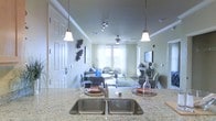 This single-floor residence showcases a thoughtfully designed open-concept layout that seamlessly connects the kitchen, dining, and living areas into one cohesive and inviting space. The kitchen is anchored by a generous granite-topped island with a double stainless steel undermount sink, elegantly lit by two pendant lights that hang above. Track lighting along the ceiling provides additional illumination, complementing the crown molding that frames the room with a refined architectural detail.

The dining area sits just beyond the kitchen island, offering a comfortable gathering space that flows naturally into the living room. The living area is bright and airy, bathed in natural light streaming through large windows dressed with sheer white curtains. A ceiling fan centered in the living space adds both function and visual balance to the room. The walls are finished in a soft, neutral tone that enhances the sense of openness throughout.

The living room is furnished with seating arranged around a light-colored area rug, and a flat-screen television is mounted on the far wall. Decorative wall art and tasteful accessories add character to the space. The overall ambiance is warm, well-lit, and welcoming, with a clean, modern aesthetic that emphasizes comfort and practicality throughout the single-floor layout.