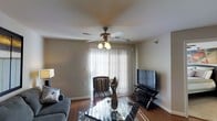 This single-floor residence offers a welcoming living area that serves as the focal point of the space. The room is anchored by warm hardwood flooring that extends throughout, adding a sense of continuity and warmth to the overall design. A ceiling fan with an integrated light fixture hangs centrally overhead, providing both air circulation and ambient lighting. Vertical blinds dress a large window or sliding door, allowing natural light to filter softly into the room while maintaining privacy. The living area is furnished with a plush sofa and an accent chair featuring a classic striped upholstery, complemented by a reflective glass coffee table that adds a modern touch. A flat-screen television is mounted on a sleek black media stand positioned along one wall, creating a practical entertainment setup. A decorative vase with dried botanicals adds a subtle natural element to the space. The neutral wall tones contribute to a clean, versatile aesthetic that accommodates a variety of décor styles. Overall, the space presents a functional and comfortable living environment with a balanced mix of practical and decorative elements.