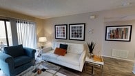 This single-floor space offers a thoughtfully designed living area that combines modern aesthetics with comfortable functionality. The living room serves as the centerpiece of the space, featuring hardwood-style flooring that adds warmth and a contemporary feel throughout. Neutral, warm-toned walls provide a versatile backdrop, complemented by a textured ceiling that adds subtle character to the room.

The living area is well-proportioned, offering ample room for seating and entertaining. Large vertical blinds along one wall suggest access to natural light and a connection to the outdoors. The room is styled with a tufted white sofa accented by colorful throw pillows, paired with a glass coffee table and eclectic decorative accessories that contribute to a modern, curated look. Two bold, framed art pieces featuring circular patterns hang prominently on the main wall, serving as a clear focal point and adding a vibrant splash of color to the otherwise neutral palette.

Additional furnishings, including a side table with glassware and a tall potted plant, enhance the livability of the space. The overall ambiance is clean, bright, and inviting, reflecting a well-maintained interior with a contemporary residential design sensibility.