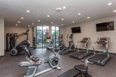 Located at 1701 Broad St in Chattanooga, Tennessee, this fitness center offers a well-equipped, single-floor exercise facility designed to support a variety of workout routines. The space is divided into distinct functional zones, each outfitted with purpose-specific equipment. The main cardio area features multiple treadmills, elliptical trainers, a StairMaster, and a rowing machine, all positioned on durable rubberized flooring. A large floor-to-ceiling window floods the space with abundant natural light and provides views of the outdoors, creating an open and energizing atmosphere. A wall-mounted flat-screen television is positioned for easy viewing during workouts. A dedicated free weights area includes a well-stocked dumbbell rack with a variety of weight options and an adjustable workout bench, ideal for strength training. Medicine balls in assorted sizes add further versatility for functional fitness exercises. The facility is accessed through glass double doors, lending a sleek, modern aesthetic to the entrance. Recessed lighting throughout ensures the space remains well-lit at all hours. The neutral-toned walls and clean, minimalist design contribute to a focused and professional workout environment, making this fitness center a comprehensive and practical amenity.