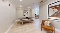Located at 3450 Miller Dr in Chamblee, GA, this thoughtfully designed single-floor space offers a versatile open layout that maximizes every square foot. The living room serves as the heart of the space, characterized by warm, neutral-toned walls, light hardwood flooring, and crisp crown molding that lend a polished, contemporary feel throughout.

A standout architectural feature is the large interior picture window that frames a dramatic view of exposed wooden truss beams beyond — a striking design detail that adds depth, character, and a connection to the building's structural artistry. Recessed lighting illuminates the space evenly, complementing the natural light that filters in through exterior windows.

The layout flows between distinct functional zones: a central gathering area anchored by a rustic wood table and bench seating, a lounge corner with a modern leather accent chair, and a built-in countertop workspace outfitted with bar-height seating — ideal for productivity or casual use. A patterned area rug defines the entry zone, adding texture and visual warmth.

An exterior door provides direct access, while a frosted-glass interior door leads to an adjacent room. The overall ambiance is clean, modern, and inviting, blending industrial accents with refined finishes to create a functional and aesthetically cohesive environment.